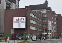 Stavba Obrtno-podjetniške zbornice Slovenije v temno bordo barvi, postavljena ob cesti; pred njo pločnik, po katerem hodijo ljudje.