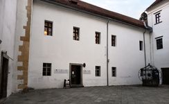 Zunanjost bele zgodovinske stavbe z rjavordečo streho in več pravokotnimi lesenimi okni; vhodna vrata obkrožajo informativne table, pred stavbo je tlakovan atrij z okrasnim kovinskim vodnjakom.